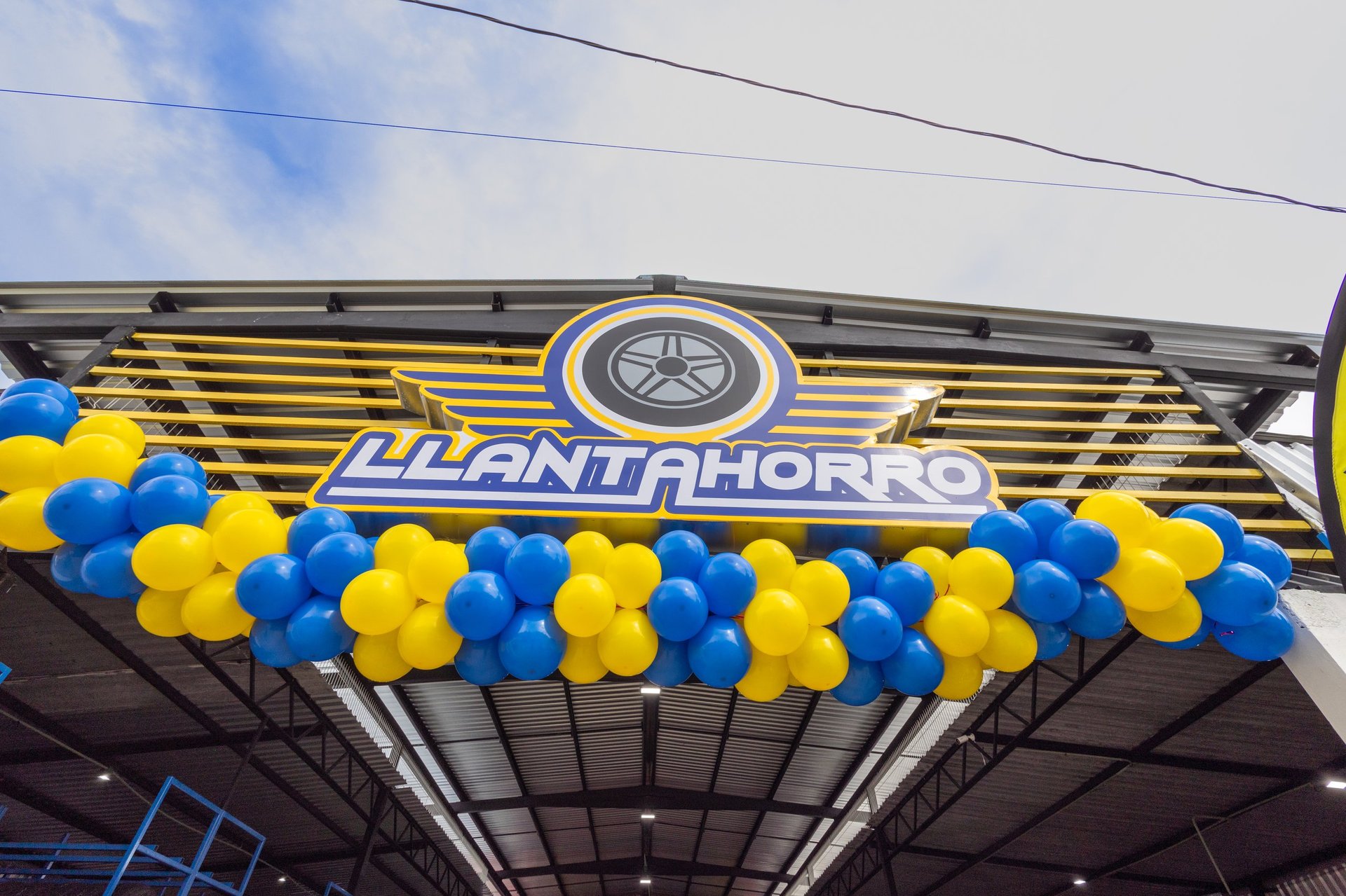 Llanthorn storefront entrance with illuminated sign, yellow and blue horizontal stripes, circular logo, and blue and yellow balloon garland decoration