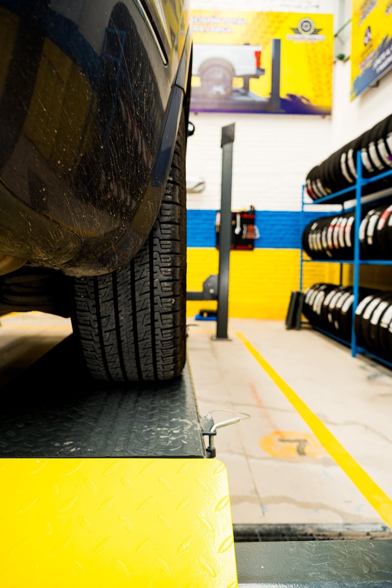 Close-up of a car tire on a lift in a tire shop with yellow and blue striped walls and tire storage racks visible in background